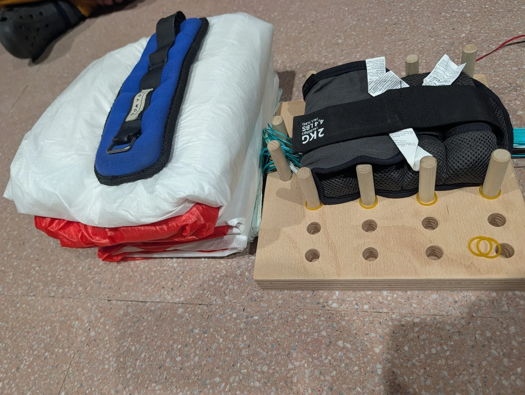 Bloque compacto de tela blanca y roja con una pesa azul encima, al lado del tablero de plegado con clavijas y gomas amarillas